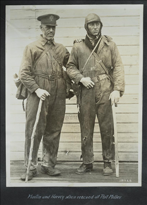 Two white men in jumpsiuts, hats, and boots, hold walking sticks. They look exhausted. 
